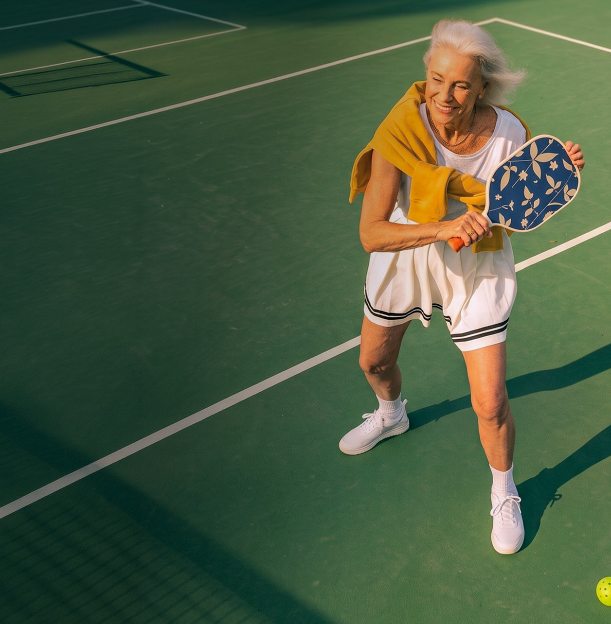 An older woman playing pickleball on a green court, wearing a white athletic dress with black trim, white sneakers, and holding a blue paddle with a yellow towel draped over her shoulder.