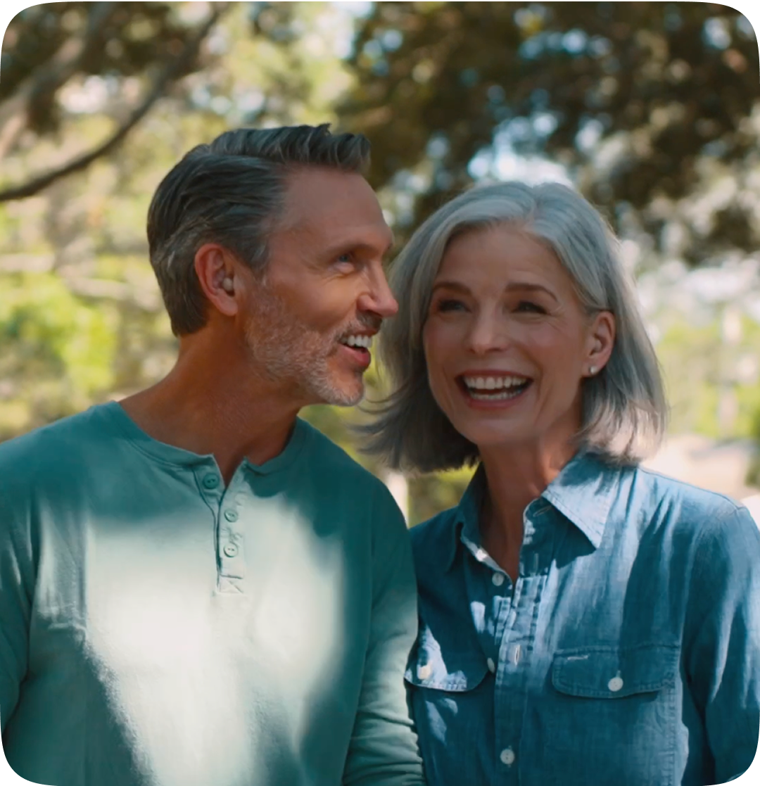 Man and woman smiling at each other outdoors with trees in the background 