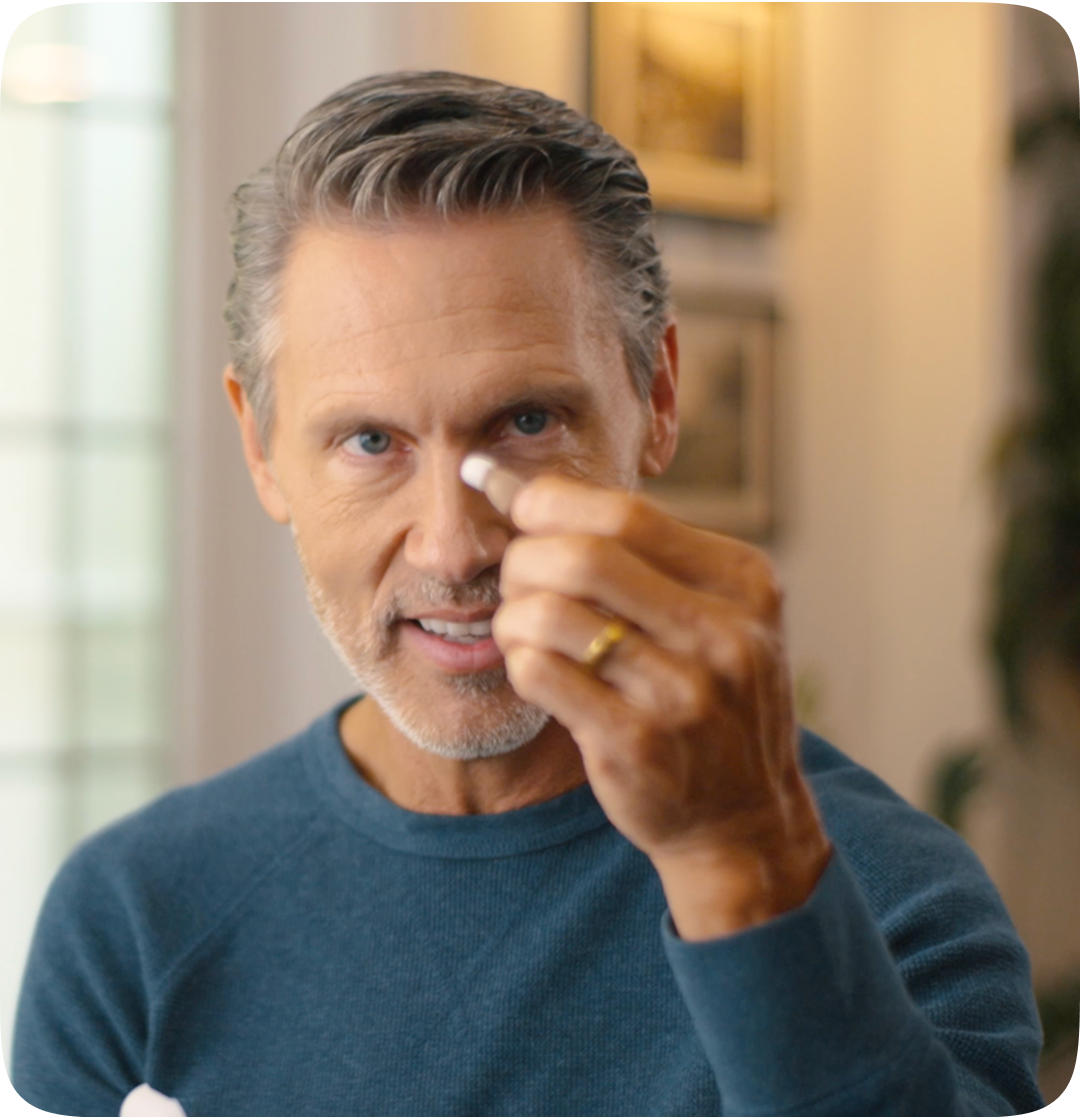 Middle-aged man in a blue sweater holding a small hearing aid between his fingers, smiling slightly while demonstrating it indoors.